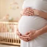 Kobieta w ciąży trzymająca delikatnie dłonie na swoim brzuchu w jasnym, przytulnym pokoju dziecięcym.