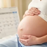 Zbliżenie na ciążowy brzuch kobiety, w tle rozmyty kalendarz. Ciepłe, naturalne oświetlenie.
