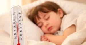 Termometr z podwyższoną temperaturą na pierwszym planie i śpiące dziecko w tle w ciepłym, przytulnym oświetleniu.