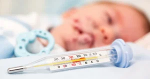 Termometr kliniczny wskazujący podwyższoną temperaturę na tle rozmytej twarzy niemowlęcia i gryzaka.