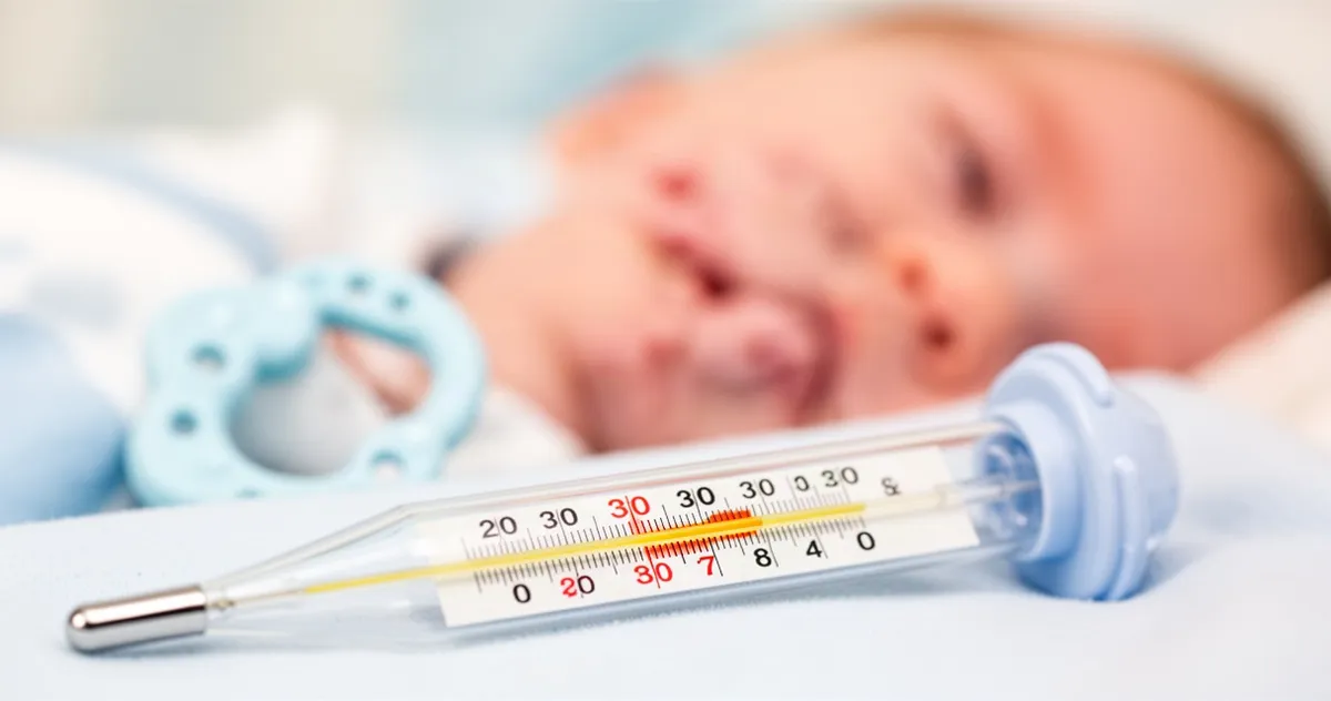 Termometr kliniczny wskazujący podwyższoną temperaturę na tle rozmytej twarzy niemowlęcia i gryzaka.