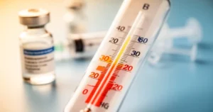 Termometr wskazujący wysoką temperaturę na tle rozmytej fiolki ze szczepionką i strzykawki w gabinecie lekarskim.