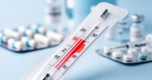 Zbliżenie na termometr wskazujący podwyższoną temperaturę, w tle rozmyte pigułki i akcesoria medyczne w odcieniach błękitu.