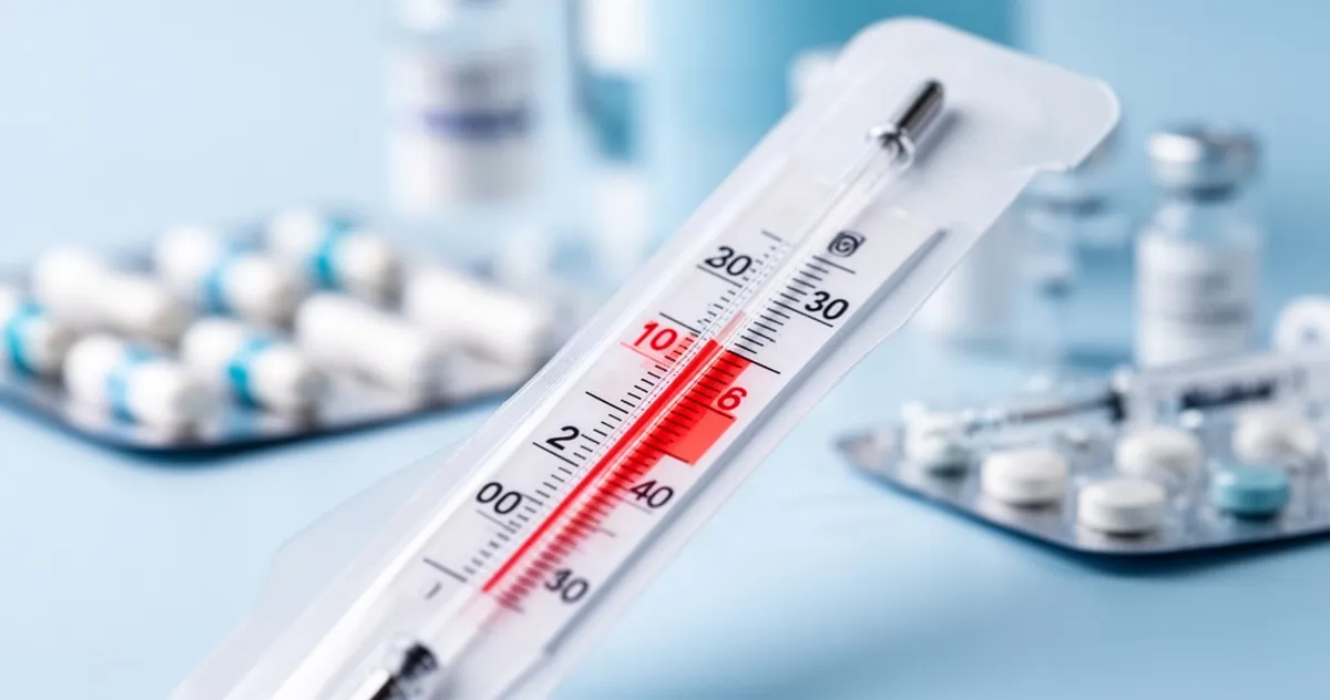 Zbliżenie na termometr wskazujący podwyższoną temperaturę, w tle rozmyte pigułki i akcesoria medyczne w odcieniach błękitu.